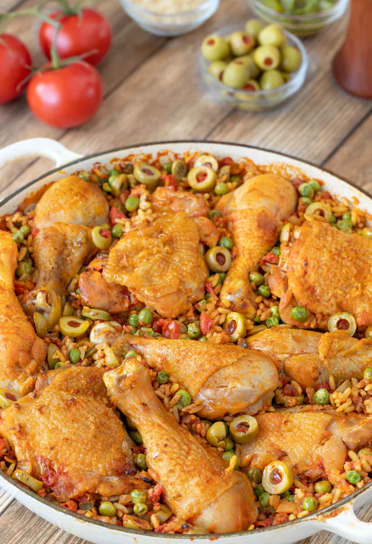 Arroz Con Pollo in a shallow Le Creuset baking dish with fresh tomatoes, stuffed green olives and other ingredients in the background