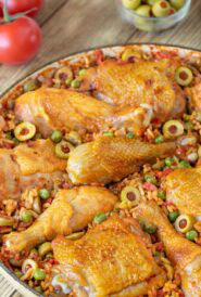Closer view of Arroz Con Pollo in a shallow Le Creuset baking dish with fresh tomatoes, stuffed green olives and other ingredients in the background