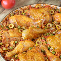 Closer view of Arroz Con Pollo in a shallow Le Creuset baking dish with fresh tomatoes, stuffed green olives and other ingredients in the background
