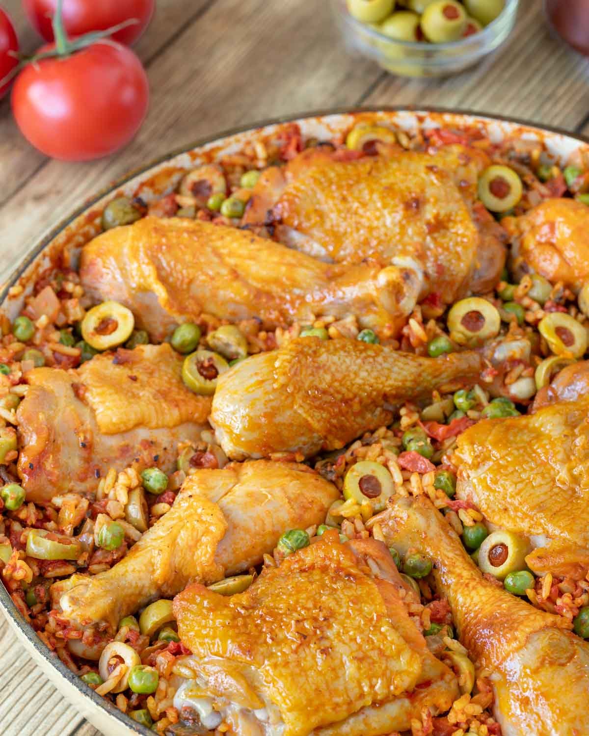 Closer view of Arroz Con Pollo in a shallow Le Creuset baking dish with fresh tomatoes, stuffed green olives and other ingredients in the background