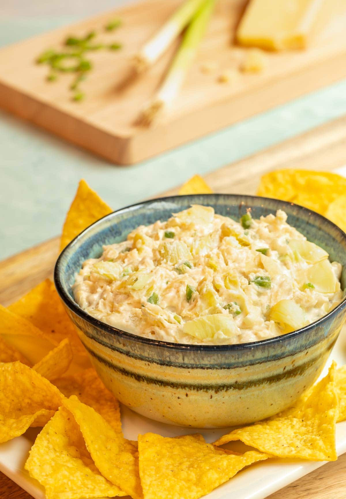 Blue striped bowl holding Jalapeno Artichoke Dip, surrouded by tortilla chips. Green onions and cheese are in the background