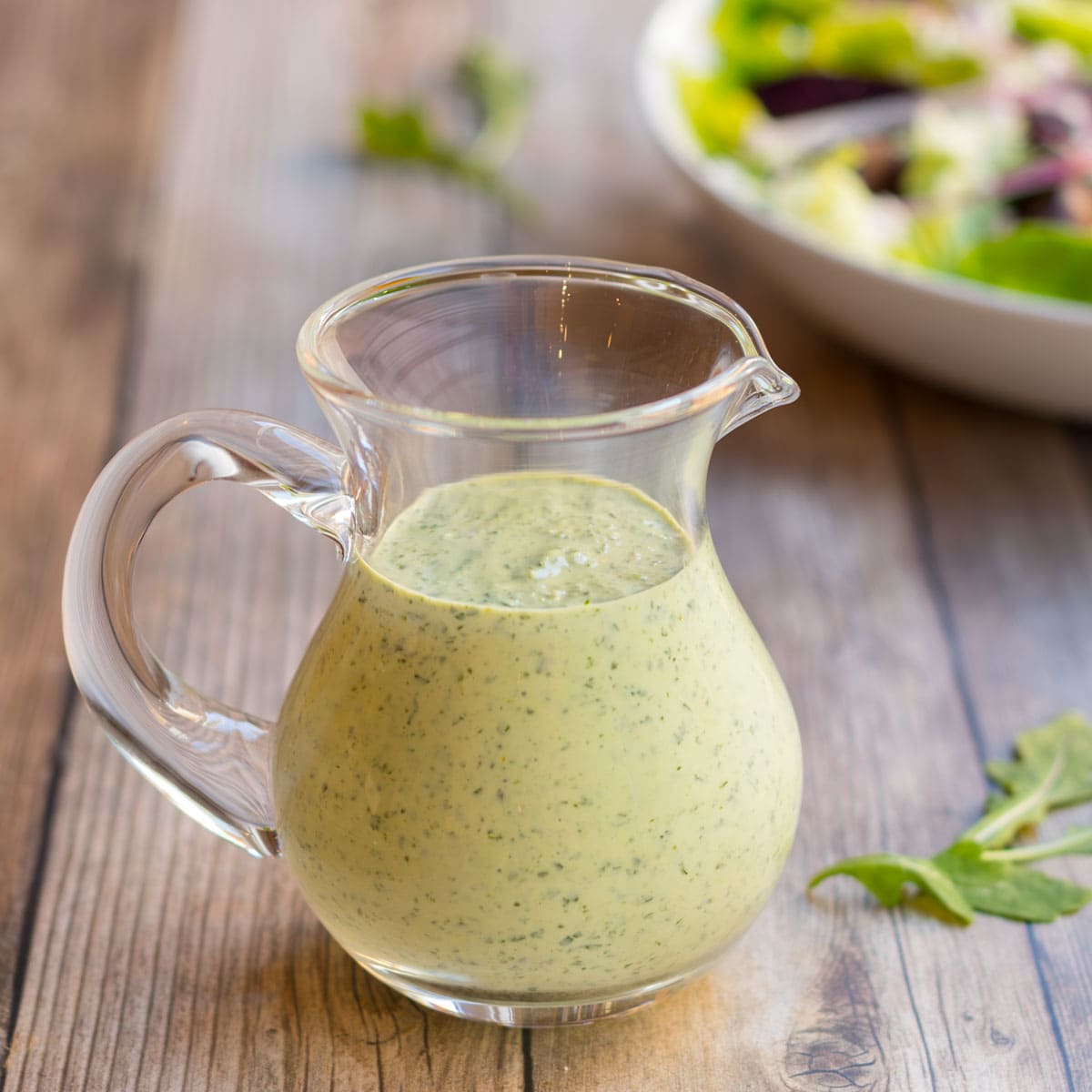 Arugula Salad Dressing in a clear glass pitcher with a salad in the background