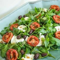 arugula spread on a blue platter, topped with roasted tomato halves, cheese and olives