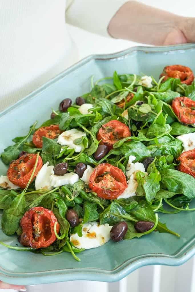 arugula spread on a blue platter, topped with roasted tomato halves, cheese and olives