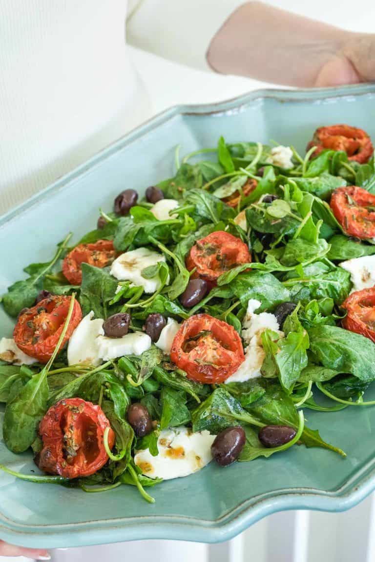 arugula spread on a blue platter, topped with roasted tomato halves, cheese and olives