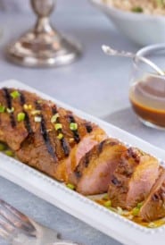 white platter filled with slices of Asian Pork Tenderloin. Bowl if rice and candles in the background