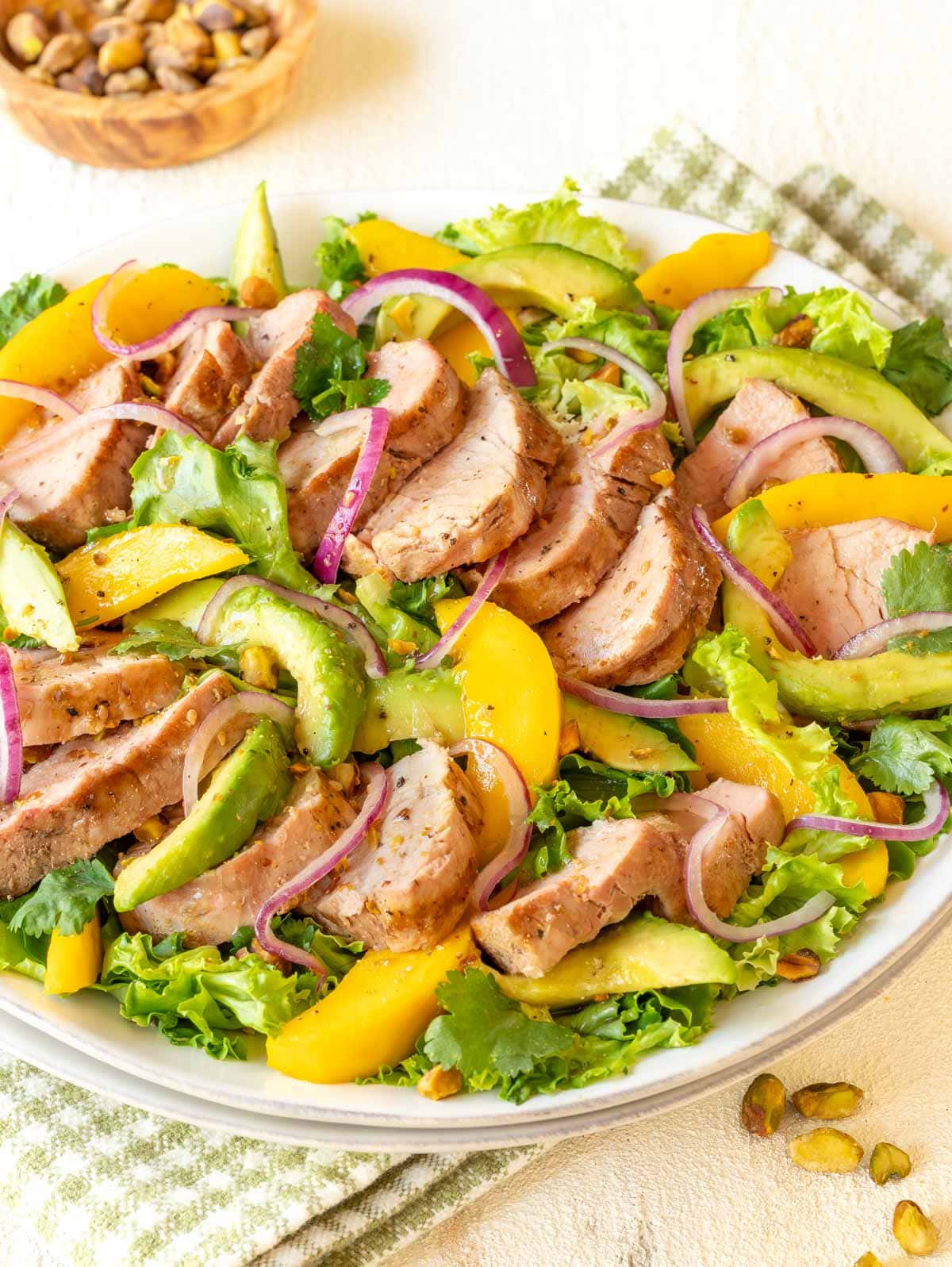 White platter holding Avocado Mango Salad with Pork Tenderloin, with bowl of pistachios in background