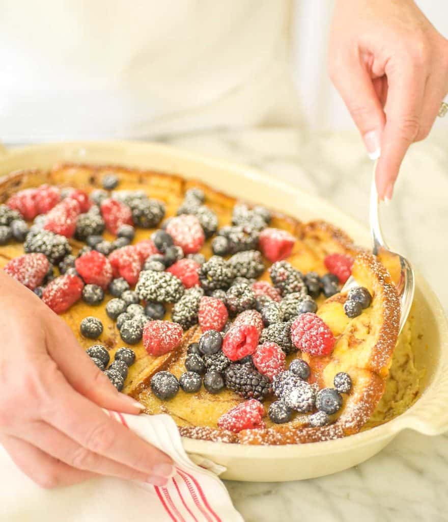 Oval baking dish filled with Baked Cinnamon French Toast, topped with berries and a spoon dishing out one serving
