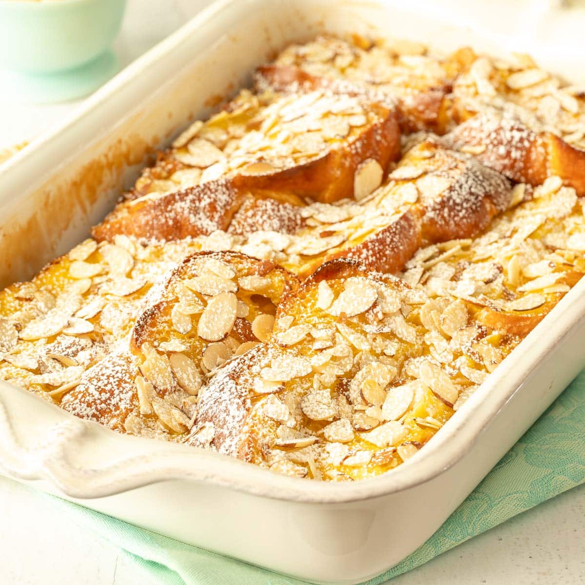 white casserole dish holding Baked Banana French Toast, with a small dish of bananas and flowers in back