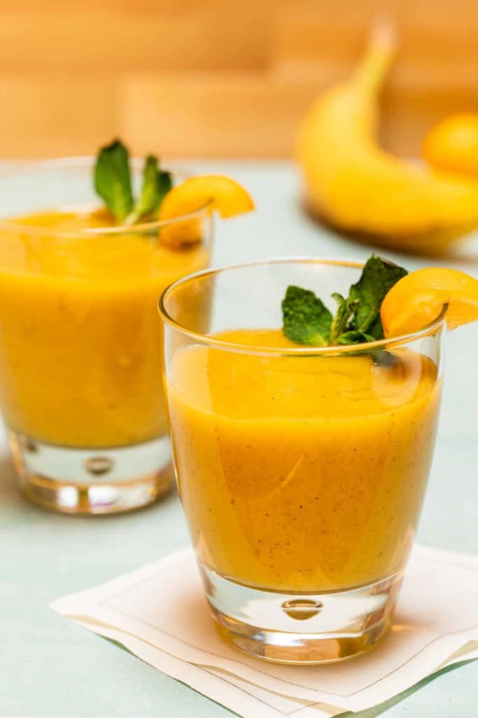 two glasses holding mango banana smoosthies with banana in the background