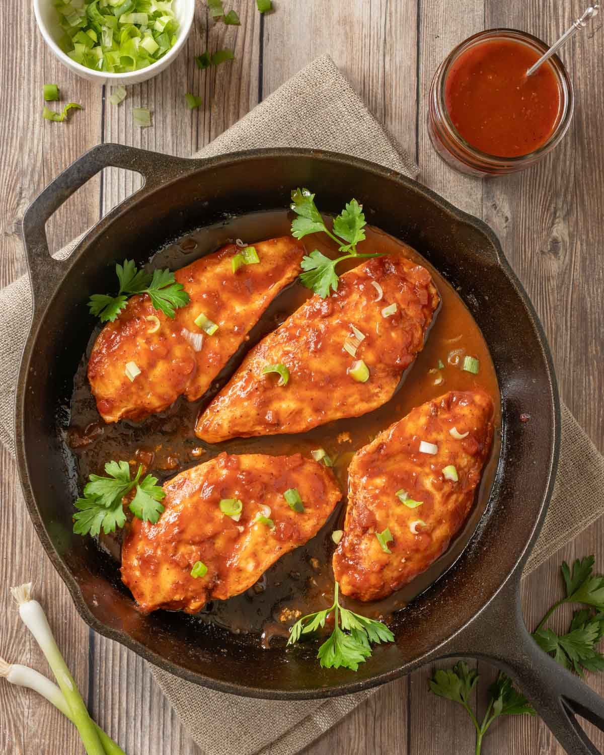 4 Baked BBQ Chicken Breasts with sauce in a cast iron skillet, with more sauce on the side