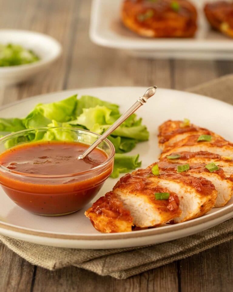white plate holding one serving of Baked BBQ Chicken Breasts with sauce in a bowl on the side and more chicken in the back