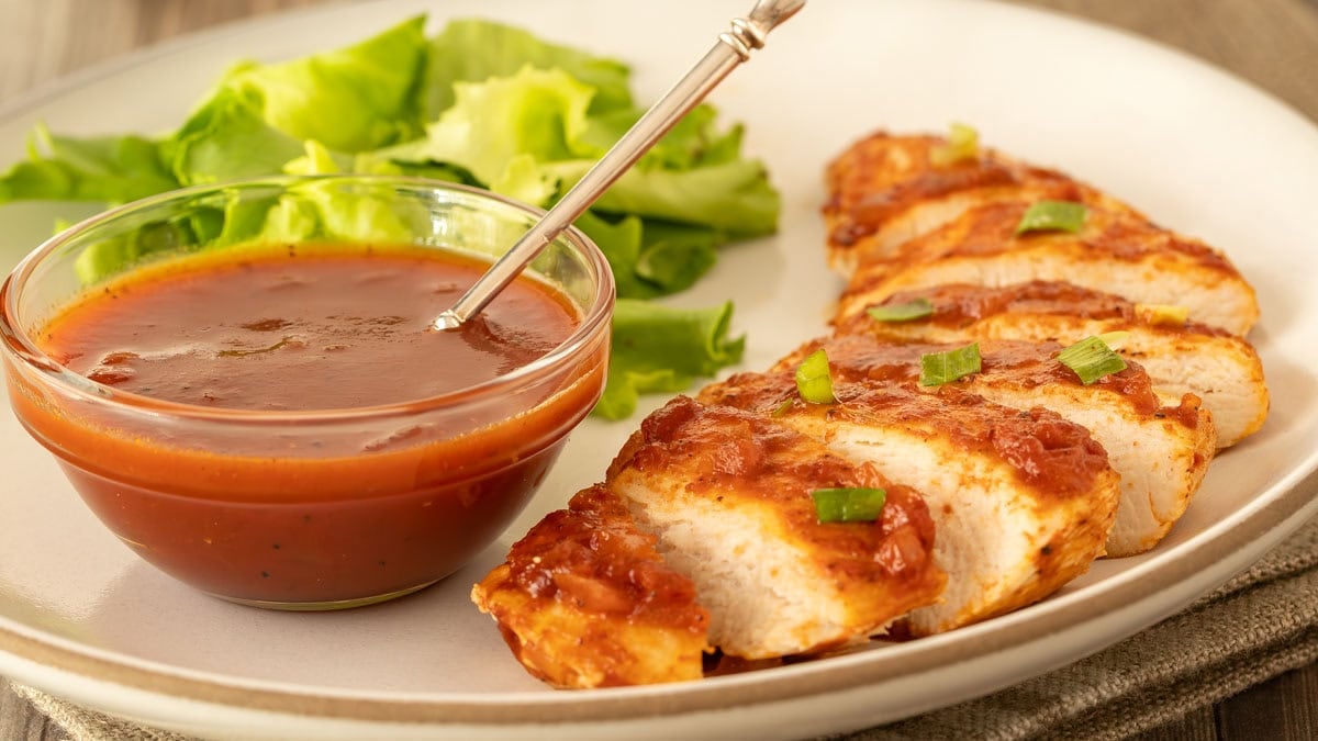 Horizontal view of a white plate holding one serving of Baked BBQ Chicken Breasts with sauce in a bowl on the side