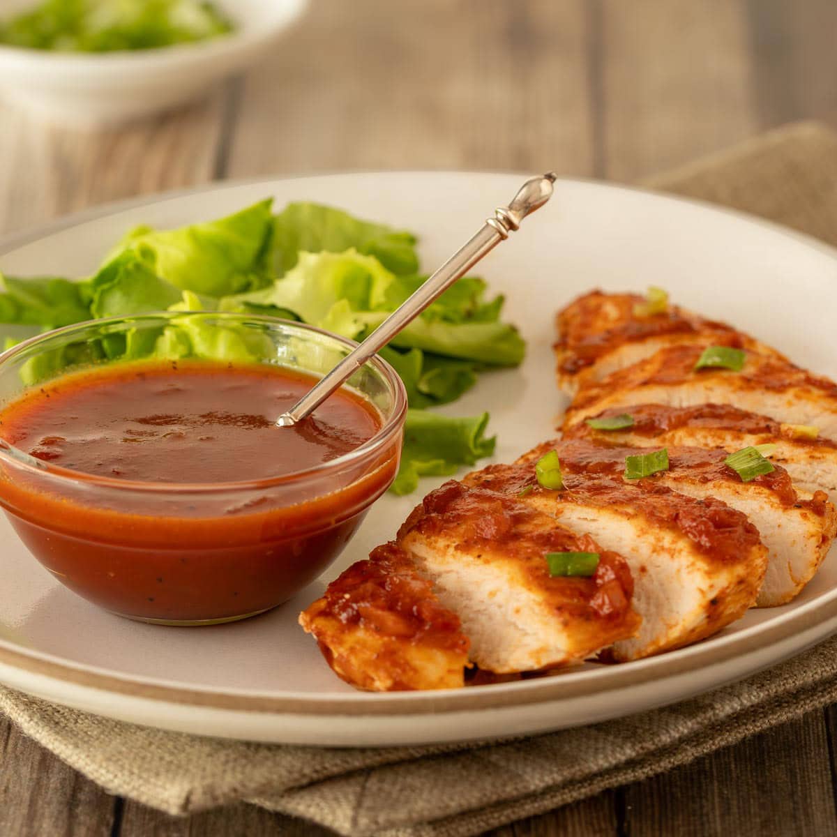 white plate holding one serving of Baked BBQ Chicken Breasts with sauce in a bowl on the side