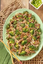 Overhead view of easy beef and broccoli stir-fry with rice noodles, served on a light green platter with wooden chop sticks and a small bowl of chopped green onions to the side.