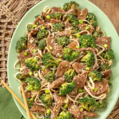 Overhead view of easy beef and broccoli stir-fry with rice noodles, served on a light green platter with wooden chop sticks and a small bowl of chopped green onions to the side.