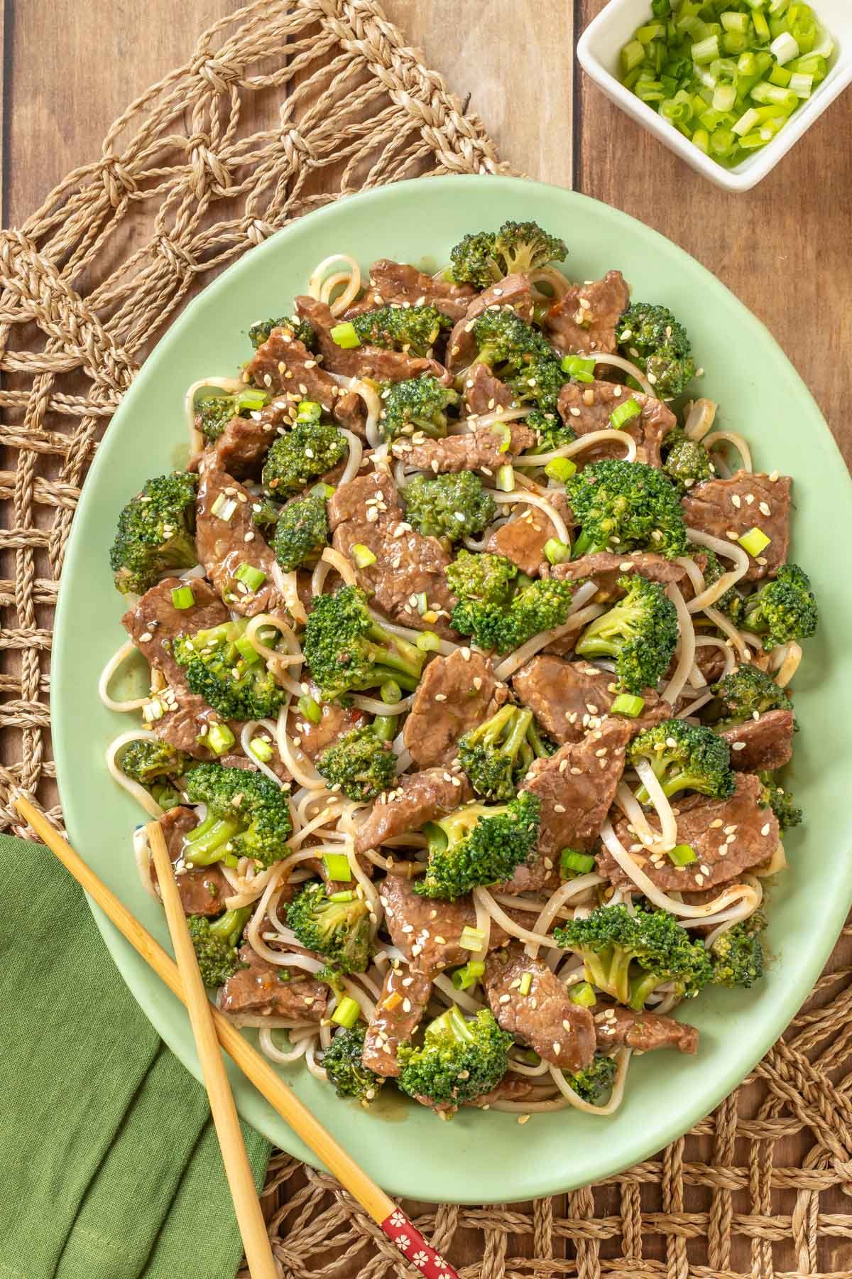 Overhead view of easy beef and broccoli stir-fry with rice noodles, served on a light green platter with wooden chop sticks and a small bowl of chopped green onions to the side.