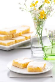 Homemade Lemon Bars stacked on a serving tray in the background, with flowers and a small plate with two lemon bars in the front