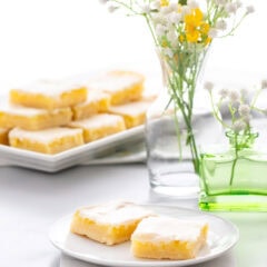 Homemade Lemon Bars stacked on a serving tray in the background, with flowers and a small plate with two lemon bars in the front