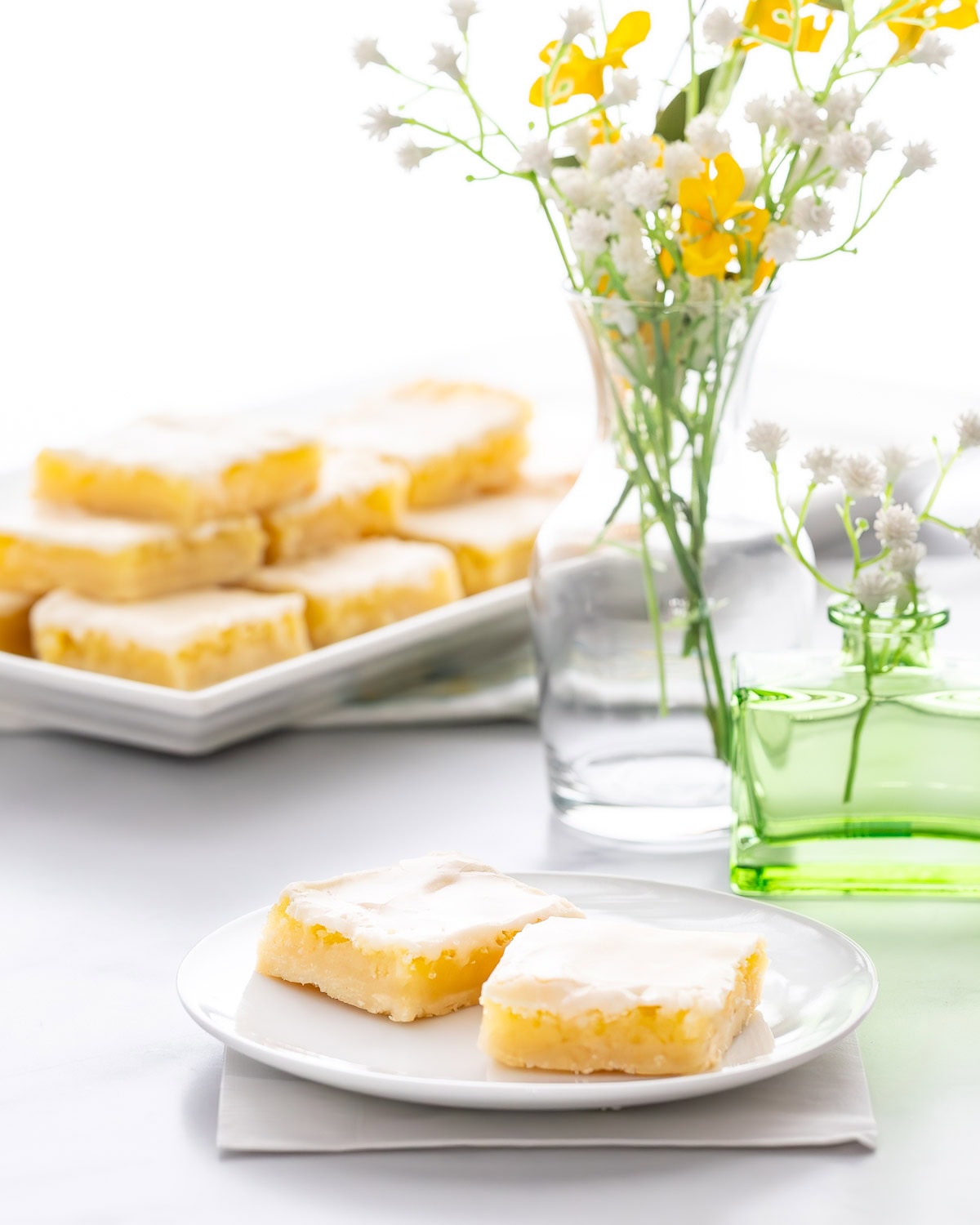 Homemade Lemon Bars stacked on a serving tray in the background, with flowers and a small plate with two lemon bars in the front