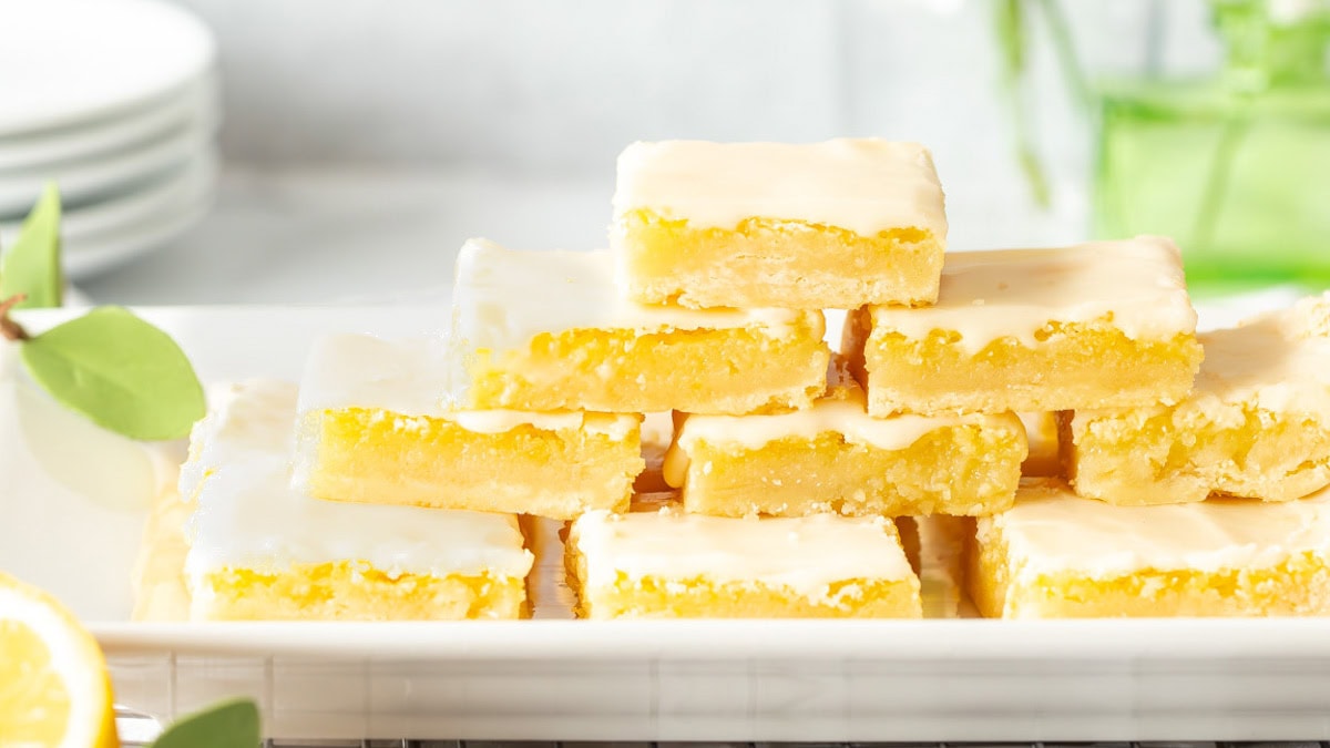 Homemade Lemon Bars stacked on a serving tray with small plates and flowers in the background
