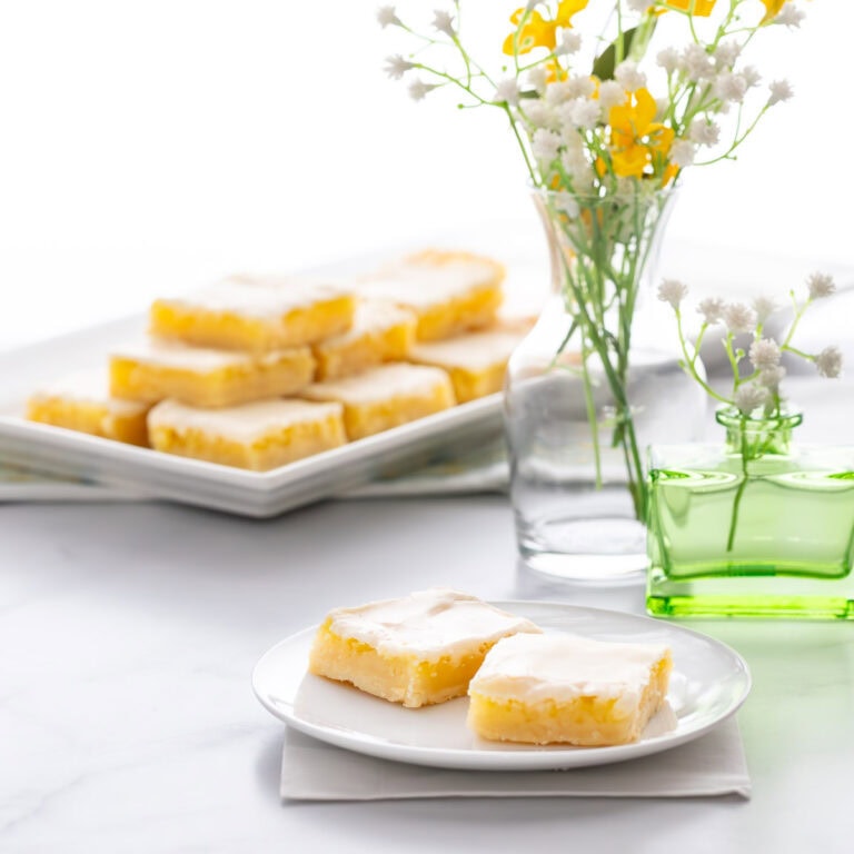 Homemade Lemon Bars stacked on a serving tray in the background, with flowers and a small plate showing two lemon bars in the front
