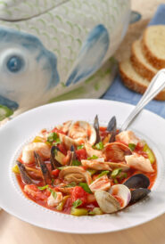 White bowl holding a portion of Bouillabaisse with a fish-shaped soup tureen and sliced bread in the background