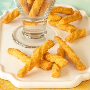 a glass holding cheese straws atop a white platter, with a few cheese straws in front