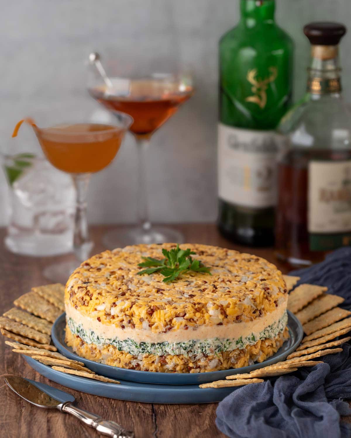 Cheddar Cheese Torte on a blue plate surrounded by crackers, with bottles of alcohol and 3 mixed cocktails in the background