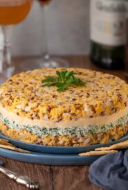 Closer view of Cheddar Cheese Torte on a blue plate surrounded by crackers, with bottles of alcohol and 3 mixed cocktails in the background