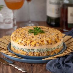 Closer view of Cheddar Cheese Torte on a blue plate surrounded by crackers, with bottles of alcohol and 3 mixed cocktails in the background