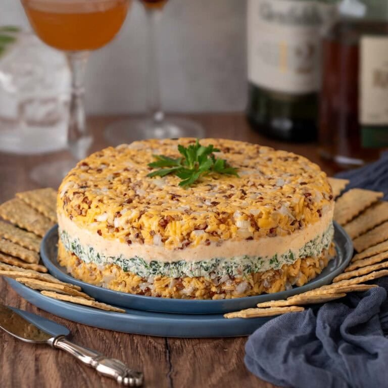 Closer view of Cheddar Cheese Torte on a blue plate surrounded by crackers, with bottles of alcohol and 3 mixed cocktails in the background