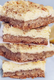 a stack of four Lemon Cheesecake Squares, with half of a lemon and a bowl of nuts in the background