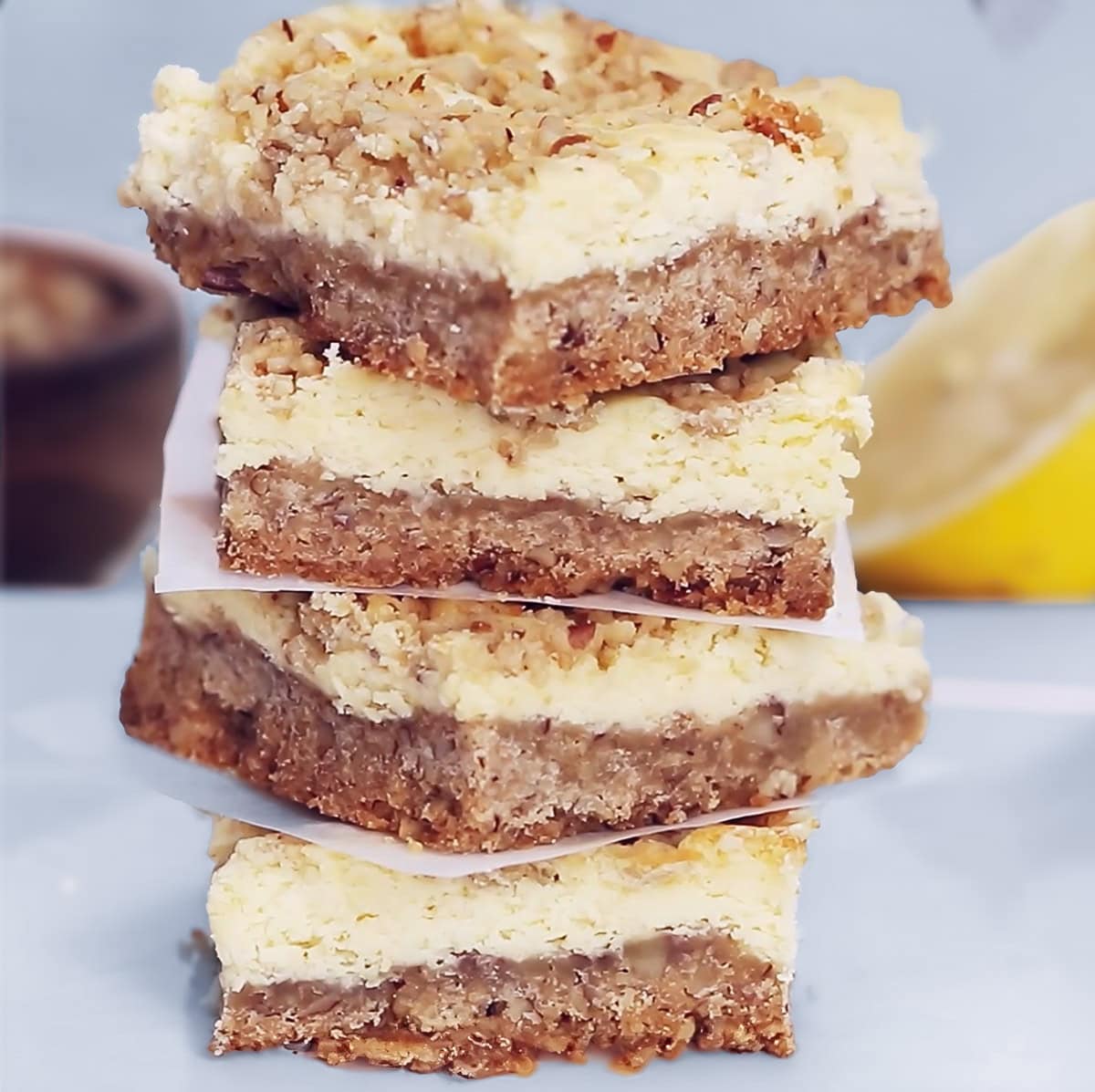 a stack of four Lemon Cheesecake Squares, with half of a lemon and a bowl of nuts in the background