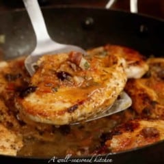 one cooked chicken breast on a metal spatula, over a pan with more chicken and tomato-olive sauce