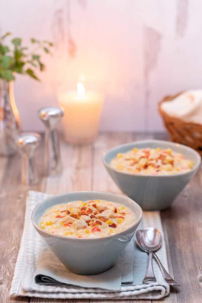 two bowls filled with Corn and Chicken Chowder with a basket of bread and a candle in the background