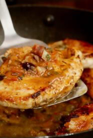 one cooked chicken breast on a metal spatula, over a pan with more chicken and tomato-olive sauce