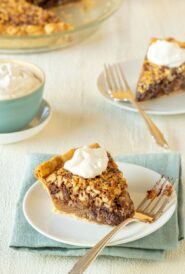 Two slices of Chocolate Pecan Pie topped with whipped cream, with a bowl of whipped cream on the side and a partial view of the whole pie in the background
