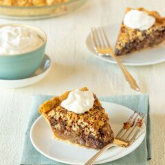 Two slices of Chocolate Pecan Pie topped with whipped cream, with a bowl of whipped cream on the side and a partial view of the whole pie in the background