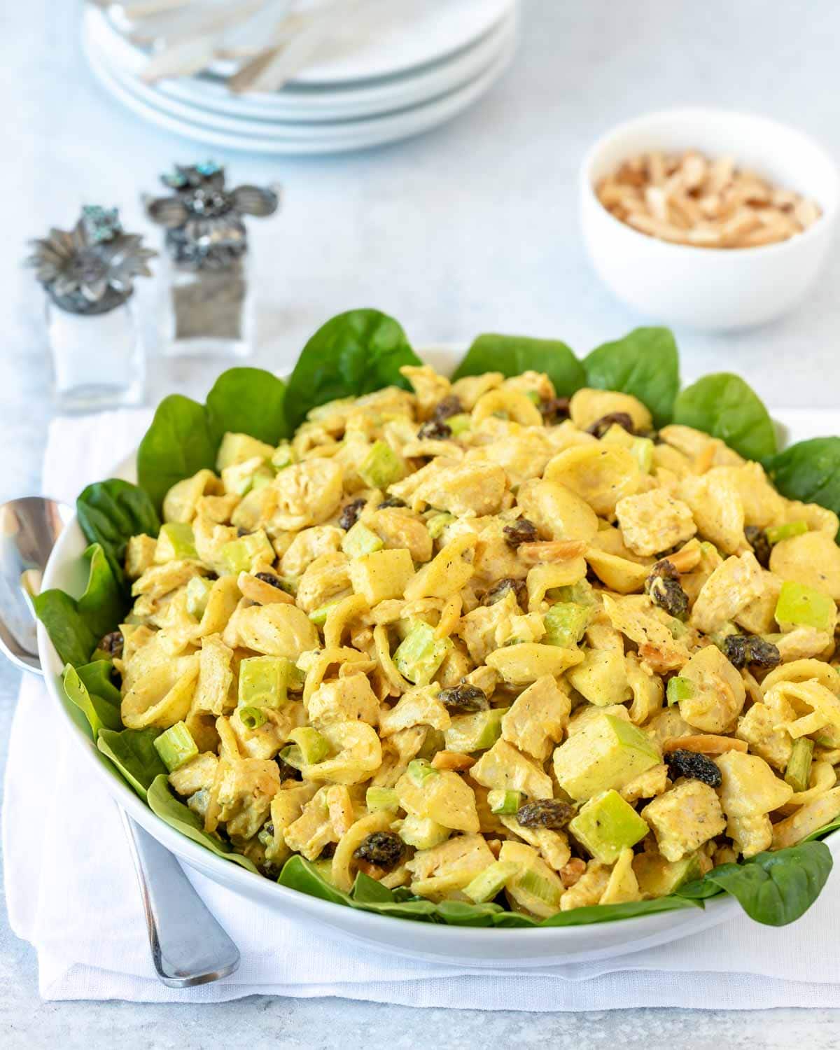 bowl lined with spinach leaves holding Curry Chicken Pasta Salad with a small bowl of almonds to the side, and a stack of plates in the background
