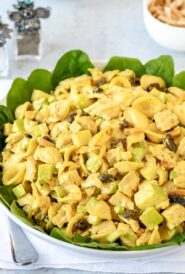 bowl lined with spinach leaves holding Curry Chicken Pasta Salad with salt, pepper, and a small bowl of almonds
