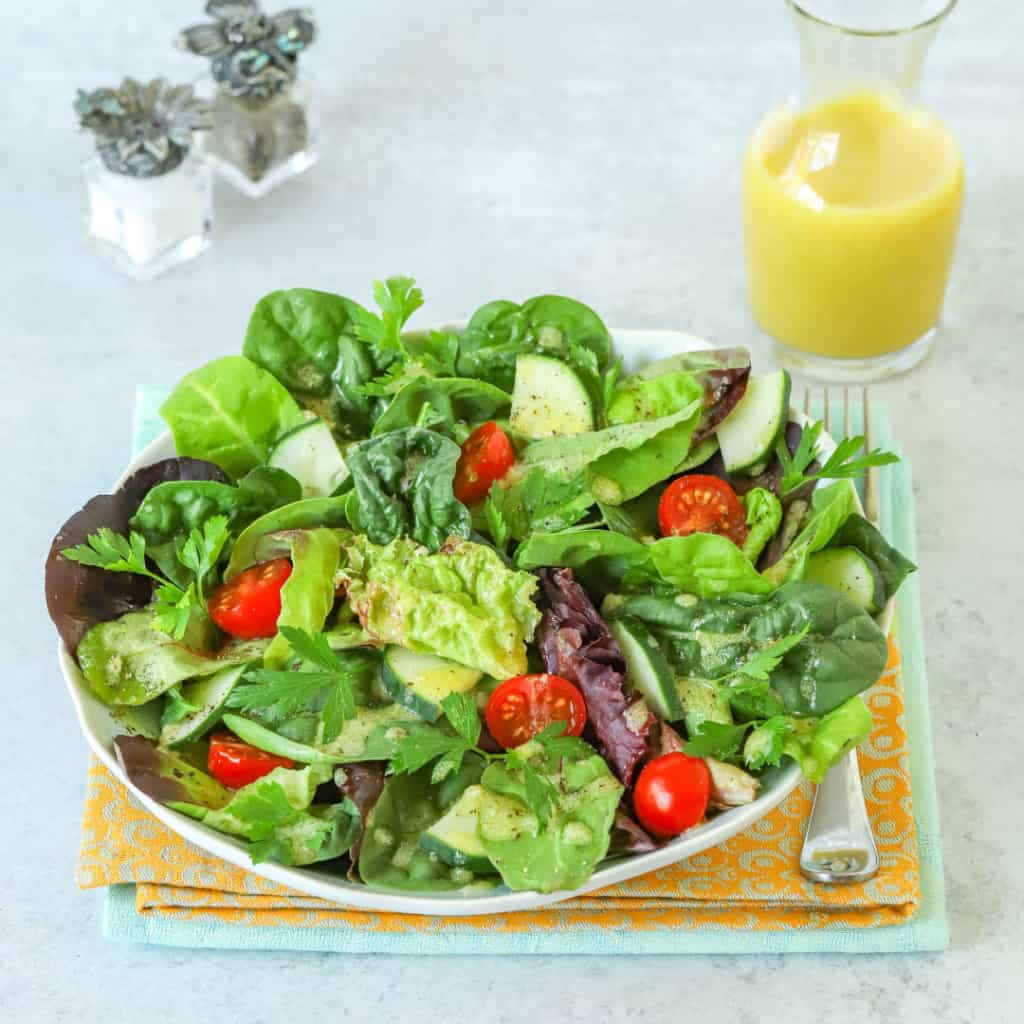Plate holding a simple mixed green salad with a carafe of Dijon Vinaigrette alongside, along with small salt and pepper shakers