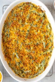 Closer overhead view of Fresh Green Bean Casserole Recipe with Cheese in a white oval baking dish, with crispy shallots and grated cheese on the side