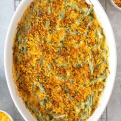 Closer overhead view of Fresh Green Bean Casserole Recipe with Cheese in a white oval baking dish, with crispy shallots and grated cheese on the side