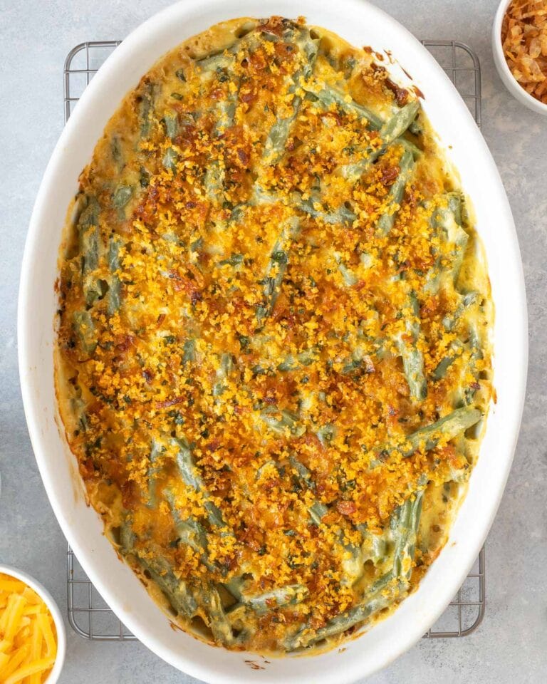 Closer overhead view of Fresh Green Bean Casserole Recipe with Cheese in a white oval baking dish, with crispy shallots and grated cheese on the side