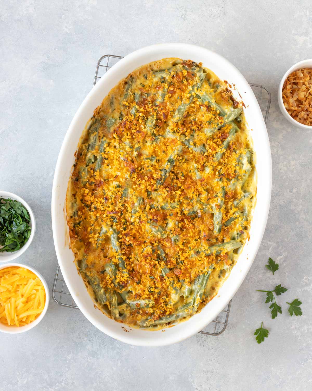 Overhead of Fresh Green Bean Casserole Recipe with Cheese in a white oval baking dish, with crispy shallots, chopped parsley and grated cheese on the side