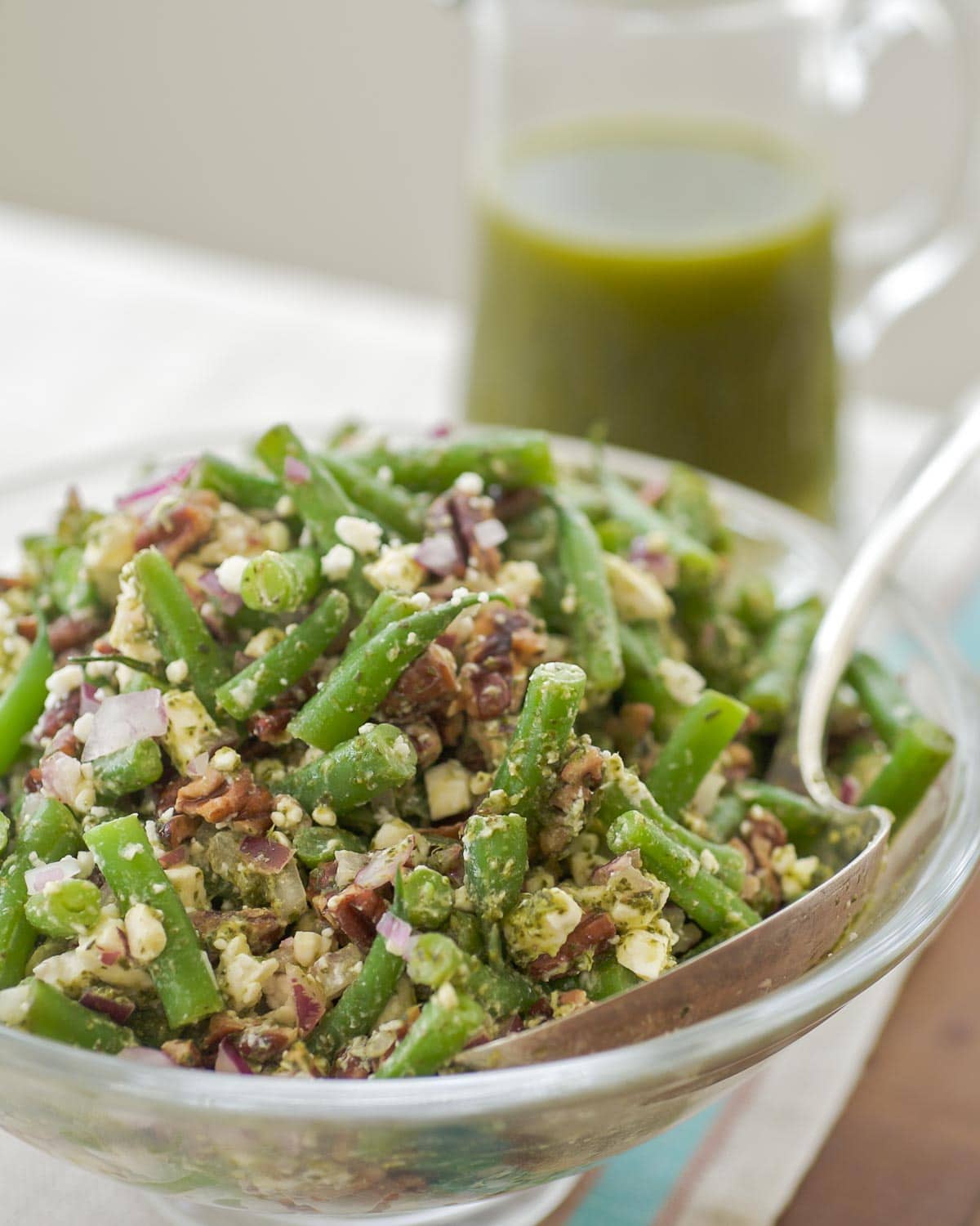 glass serving bowl holding green bean salad with mint vinaigrette in a glass pitcher in the back