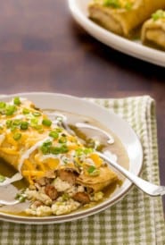 plate holding one breakfast burrito smothered in green chile sauce, with a corner open and two more on a platter in the backgrond