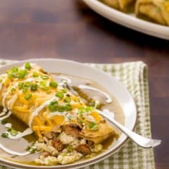 plate holding one breakfast burrito smothered in green chile sauce, with a corner open and two more on a platter in the backgrond
