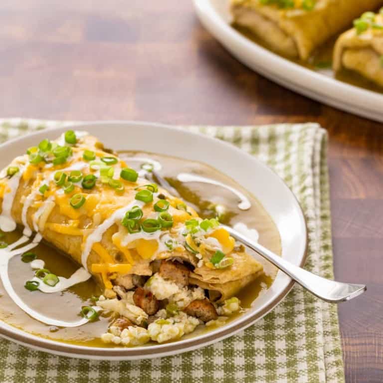 plate holding one breakfast burrito smothered in green chile sauce, with a corner open and two more on a platter in the backgrond